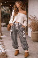 Load image into Gallery viewer, Woman wearing a white off-shoulder sweater and striped pants with brown boots, standing outdoors.
